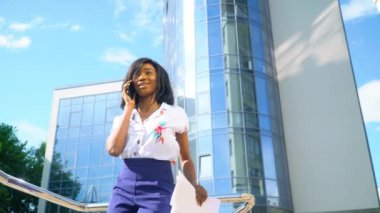 Yetişkin Afrikalı Amerikalı genç iş kadını modern ofis binasının yakınında telefonla konuşuyor.