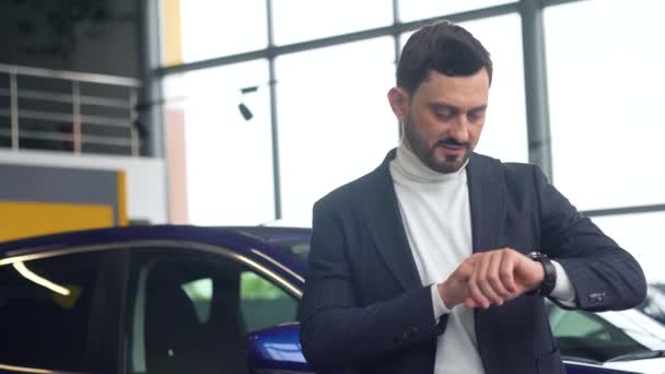 Homem elegante usa um relógio inteligente no showroom do carro. Auto negócio, venda de carros, tecnologia e conceito de pessoas — Vídeo de Stock