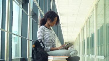 Yerde oturan Afrikalı Amerikalı kadın öğrenci üniversitede bir tablet kullanıyor. Yeni modern tam işlevsel eğitim tesisi. Çevrimiçi eğitim kavramı