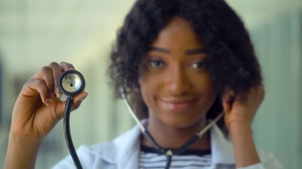 Amigável feliz Africano americano mulher médico ou enfermeiro profissional clínico geral posando com estetoscópio olhando para a câmera no consultório médico. Conceito de medicina, cuidados de saúde e pessoas — Vídeo de Stock