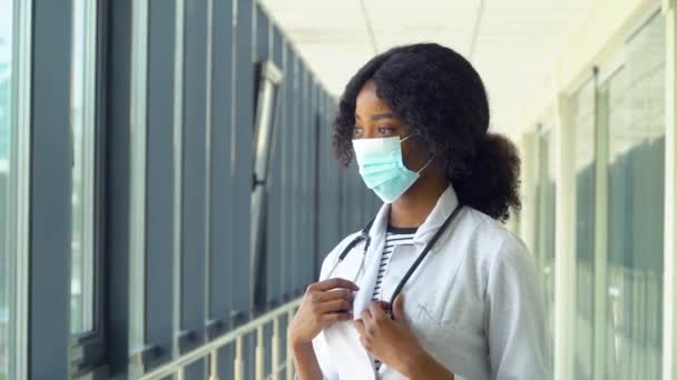 Médico o enfermera afroamericana posando con estetoscopio en máscara protectora mirando la cámara en el consultorio médico. Concepto de medicina, salud y personas — Vídeos de Stock