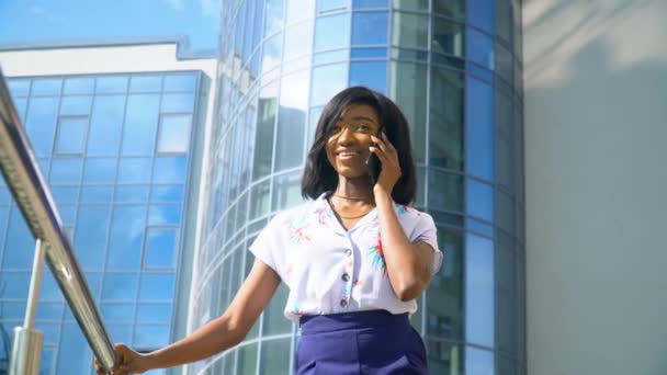 Volwassen Afro-Amerikaanse jonge zakenvrouw praat over de telefoon buiten in de buurt van modern kantoorgebouw — Stockvideo