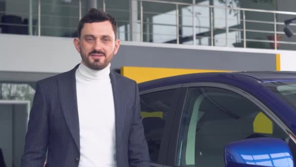 Homem elegante está olhando para a câmara de pé perto do carro azul no showroom do carro. Conceito de tecnologia e pessoas — Vídeo de Stock