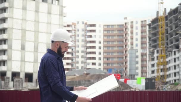 Portret van een succesvolle jonge ingenieur, architect, bouwer, zakenman, met een witte helm op, in een shirt, met een project in zijn hand, een wolkenkrabber achtergrond en een bouwplaats — Stockvideo