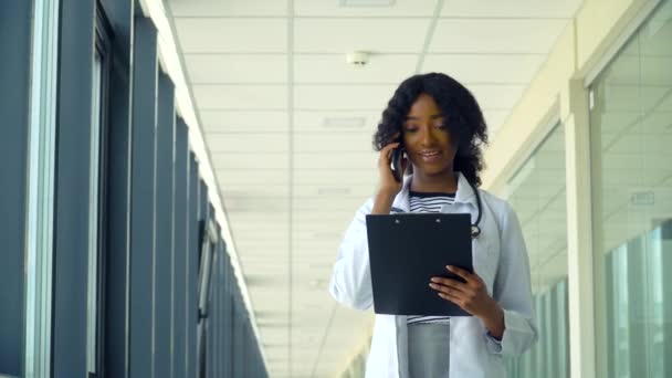 在一家现代化医院的走廊里，一位非洲裔美国护士拿着笔记本在电话上交谈，笑着。医药、保健和人的概念 — 图库视频影像