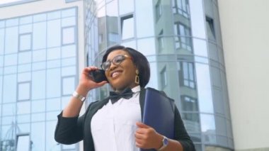 Takım elbiseli ve beyaz gömlekli Afro-Amerikalı genç iş kadını modern ofis binasının önünde telefonla konuşuyor. Karantinanın sonu