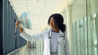 Kadın Afro-Amerikalı doktor, ciğerlerinin röntgenini inceleyip içeride tutuyor. Uzman, göğsünün şeffaf görüntüsünü kollarında tutuyor ve dikkatlice araştırıyor.