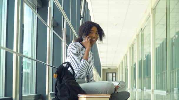 Estudiante afroamericana sentada en el piso hablando por teléfono en el pasillo de la universidad. Nueva instalación educativa moderna totalmente funcional. Concepto de educación en línea — Vídeos de Stock