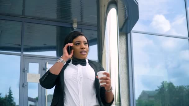 Afro-Amerikaanse jonge zakenvrouw in pak en wit shirt praat over de telefoon buiten in de buurt van modern kantoorgebouw — Stockvideo