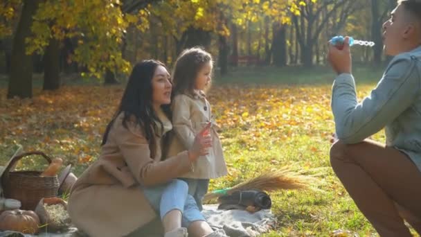 Aile hava kabarcıkları üflüyor. Anne ve baba ve kızı dışarıda eğleniyorlar. Sonbahar parkında piknik — Stok video