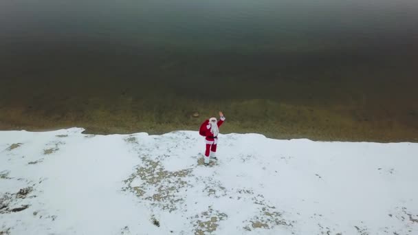 サンタクロースは湖の雪の海岸にギフトバッグで立っています。北極圏のサンタ。トップ表示 — ストック動画