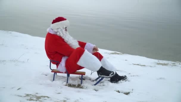 サンタクロースは海の雪の海岸にそりに座って、水の近くのそりで前進しようとします。海のサンタ — ストック動画