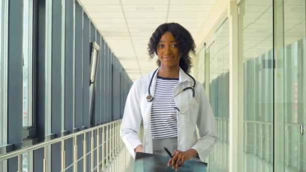 Porträt einer lächelnden afrikanisch-amerikanischen Ärztin, die mit ihrer Arbeit auf einem Flur eines modernen Krankenhauses zufrieden ist. Konzept der Medizin, des Gesundheitswesens und der Menschen — Stockvideo