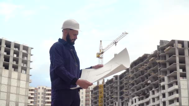 Retrato de um jovem engenheiro de sucesso, arquiteto, construtor, empresário, vestindo um capacete branco, em uma camisa, segurando um projeto em sua mão, um fundo arranha-céu e um canteiro de obras — Vídeo de Stock