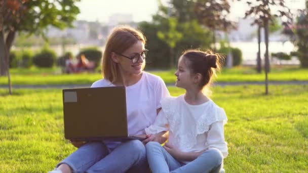 Młoda kobieta z córką pracująca jako wolny strzelec na laptopie w parku w słoneczny letni dzień. Mała dziewczynka uczy się online — Wideo stockowe