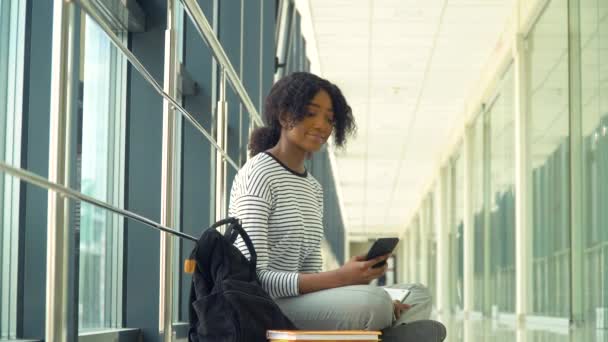 Afroamerykańska studentka siedząca na podłodze z telefonem na uniwersytecie. Nowa nowoczesna, w pełni funkcjonalna placówka edukacyjna. Pojęcie edukacji internetowej — Wideo stockowe