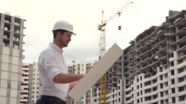 Başarılı genç bir mühendisin portresi, mimar, müteahhit, iş adamı, beyaz kasklı, gömlekli, elinde bir proje, bir gökdelen arka planı ve bir inşaat alanı tutan