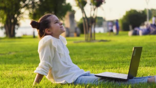 Маленька дівчинка з ноутбуком відпочиває в парку на заході сонця. Дівчина вивчає онлайн — стокове відео