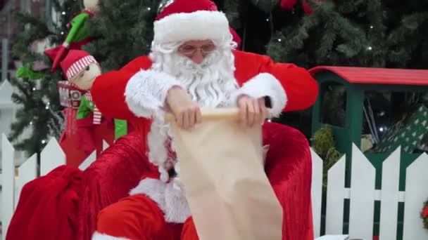 Santa Claus se sienta con una bolsa de regalos en el centro comercial escribe algo a la lista — Vídeo de stock