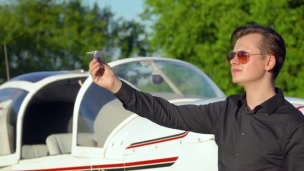 El hombre sostiene el avión de papel como en la infancia delante del avión pequeño estacionado. Pequeño concepto de aviación — Vídeo de stock