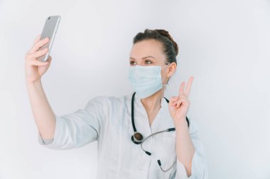 Koruyucu maskeli genç hemşire beyaz bir arka planda selfie çekiyor.