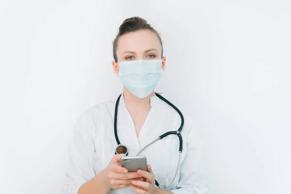 Jovem enfermeira em uma máscara protetora usa seu smartphone em um fundo branco em um hospital — Fotografia de Stock