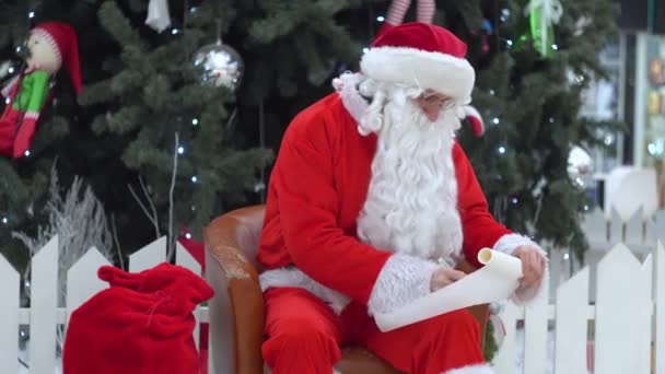 サンタクロースはショッピングモールにプレゼントの袋と一緒に座ってリストに何かを書きます — ストック動画