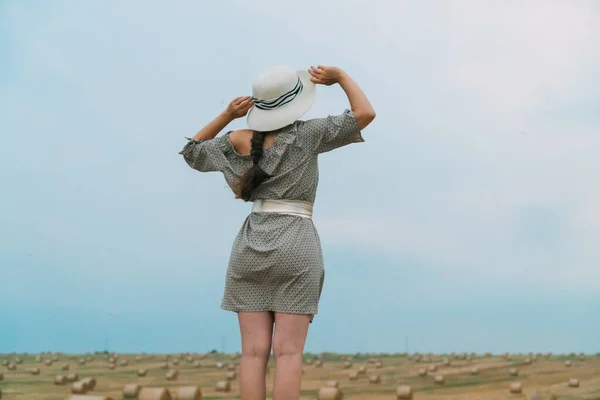 Menina bonita em um chapéu olha para o céu no meio de um campo de trigo com fardos na noite de verão — Fotografia de Stock