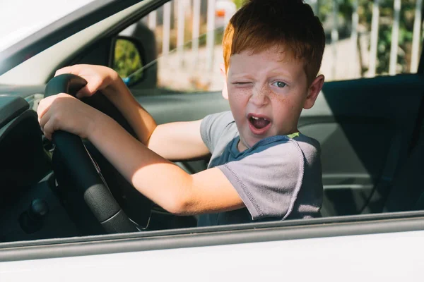 Cute little red-haired boy sitting in front of the car holding steering wheel. Boy looking at the camera and smiling — Stock Photo, Image