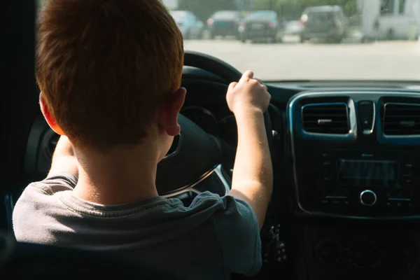 Pequeño chico pelirrojo se sienta delante del coche sosteniendo el volante, vista trasera — Foto de Stock