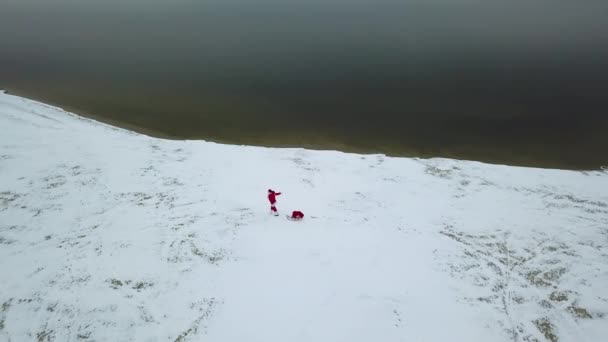 Santa Claus tira de un trineo con una bolsa de regalos a lo largo de la costa nevada del océano. Vista superior — Vídeos de Stock