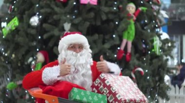 Noel Baba kameraya iyi bir jest yapıyor. Noel Baba alışveriş arabasının arkasında bir hediyeyle saklanıyor.