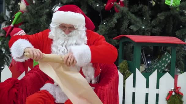 Santa Claus se sienta con una bolsa de regalos en el supermercado escribe algo a la lista — Vídeo de stock