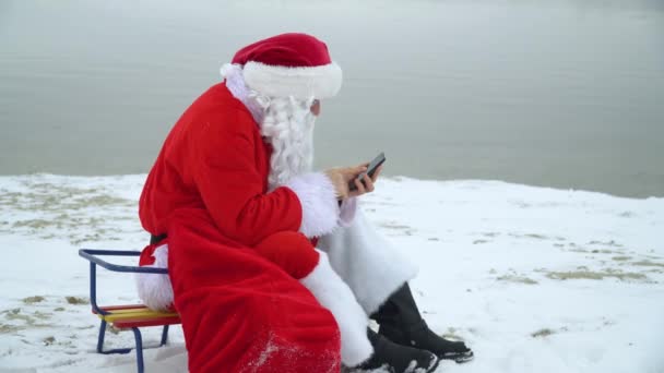 Санта Клаус сидит с мешком подарков на снежном берегу океана и работает над своим смартфоном — стоковое видео