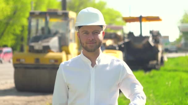 Portret van de manager van de reparatiewerkzaamheden in een witte helm tegen de achtergrond van een wegenbouwplaats — Stockvideo