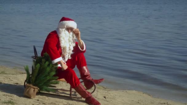 De Kerstman zit in een slee op de zandoever van het meer, repareert zijn baard, bril en pet — Stockvideo