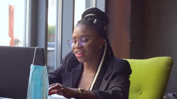 Mujer joven afroamericana comunicándose por conferencia telefónica hablar mirando la computadora en la cafetería. Máscara médica desechable colgando en la pantalla. Trabajo durante el autoaislamiento y la cuarentena — Vídeo de stock