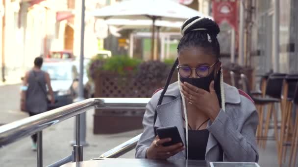 Αφροαμερικανή κοπέλα με μάσκα συνομιλεί με φίλους και πίνει καφέ στο street cafe — Αρχείο Βίντεο