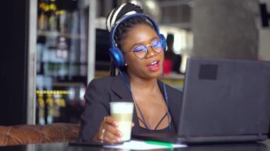 Genç siyahi kadın satış temsilcisi kulaklıkla müşteriyle konuşuyor. Video konferans görüşmesi yapıyor. Laptop içeceklerine bakıyor.
