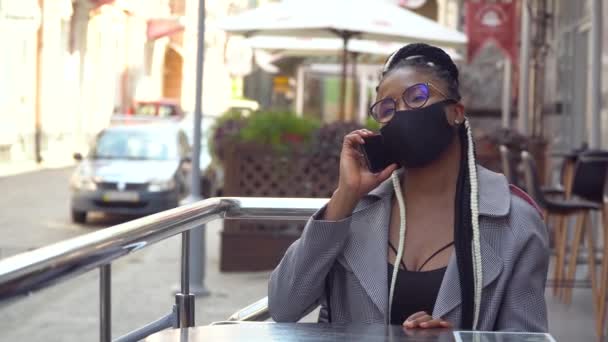 Joven mujer negra con máscara habla por teléfono en la cafetería de la calle. Hermosa mujer usando el teléfono móvil para hablar — Vídeo de stock