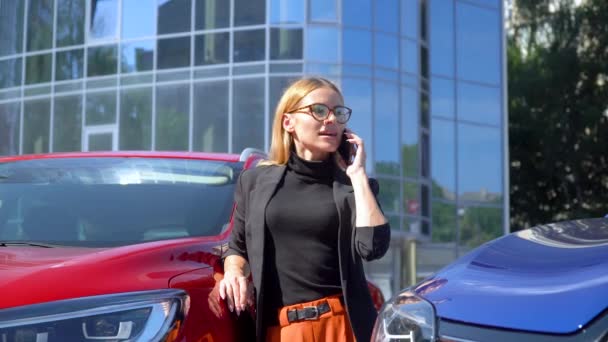 Pomyślne businesswoman stoją w pobliżu luksusowych samochodów podczas rozmowy na smartfonie w pobliżu centrum biznesowego — Wideo stockowe