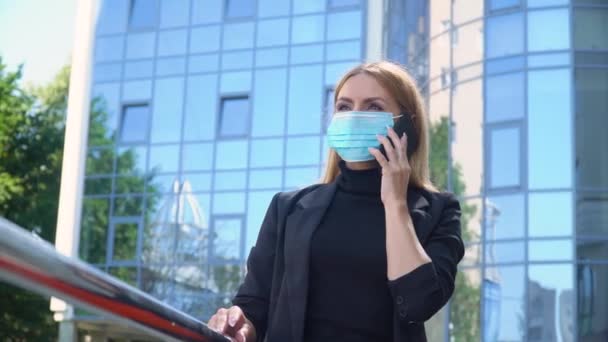 マスクの若いビジネスマンの女性は、街の通りに立って、美しいブロンドの夏の日に電話で話している呼び出しを行っている。そうだ。社会的距離の注意事項で仕事に戻る — ストック動画