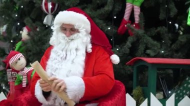 Noel Baba elinde bir torba hediyeyle süpermarkette oturuyor. Listeye bir şeyler yazıyor.