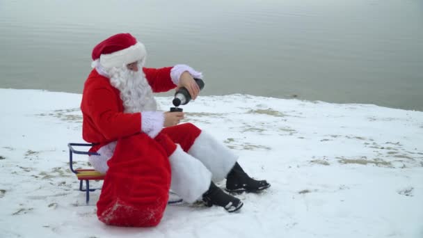 Jultomten sitter med en påse presenter på den snöiga stranden av en sjö och dricker kaffe från en termos — Stockvideo