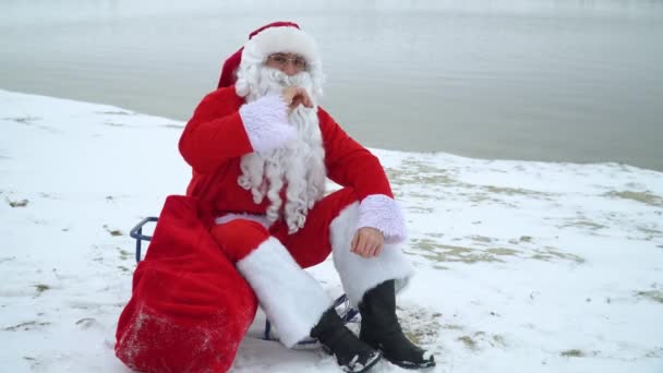 Babbo Natale si siede con un sacchetto di regali sulla riva innevata di un lago. Babbo Natale gesti OK per la fotocamera — Video Stock