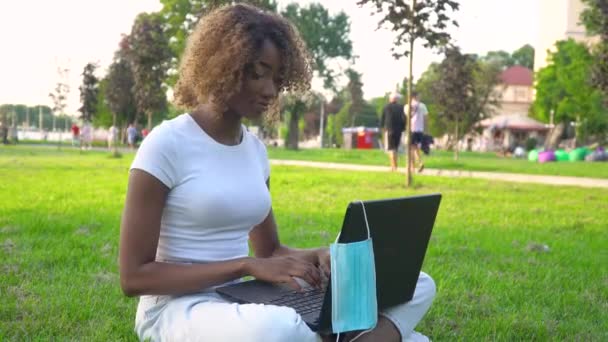 Ung afrikansk amerikansk kvinna använder laptop i parken under covid 19 pandemi. Medicinsk engångsmask hängande på displayen — Stockvideo