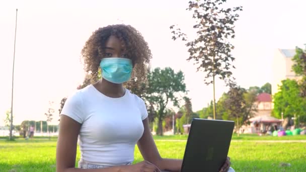 Joven estudiante afroamericana en máscara protectora trabajando en laptop en parque durante covid 19 pandemia — Vídeo de stock