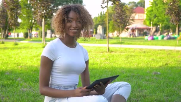 Gyönyörű mosolygós magabiztos fiatal afro-amerikai nő csinos arc tabletta nézi kamera pózol egyedül a parkban — Stock videók