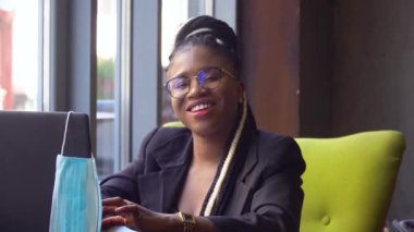 Genç siyah kadın dizüstü bilgisayar kullanıyor ve kameraya bakıyor. Ev ofisinden bağımsız çalışma, kendini izole etme. Ekranda tek kullanımlık tıbbi maske asılı. Coronavirus salgını Covid 19