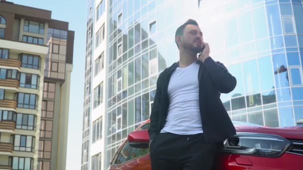 Молодий бізнесмен стоїть біля машини під час розмови на смартфоні біля бізнес-центру — стокове відео